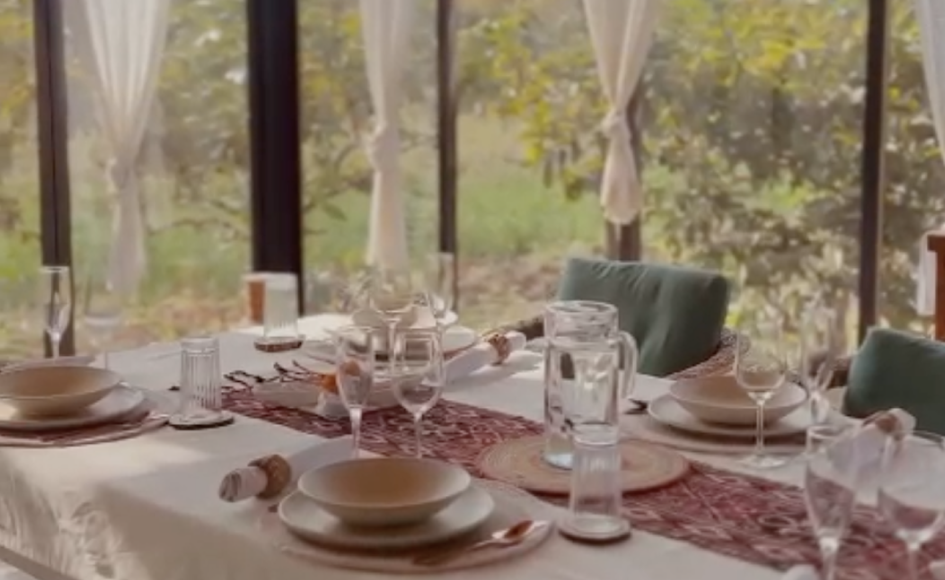 Elegant dining table setup by a sunlit glass window, featuring neutral tableware, wine glasses, and cozy green cushions only at Dhyaana Farms the boutique hotel in Aurangabad