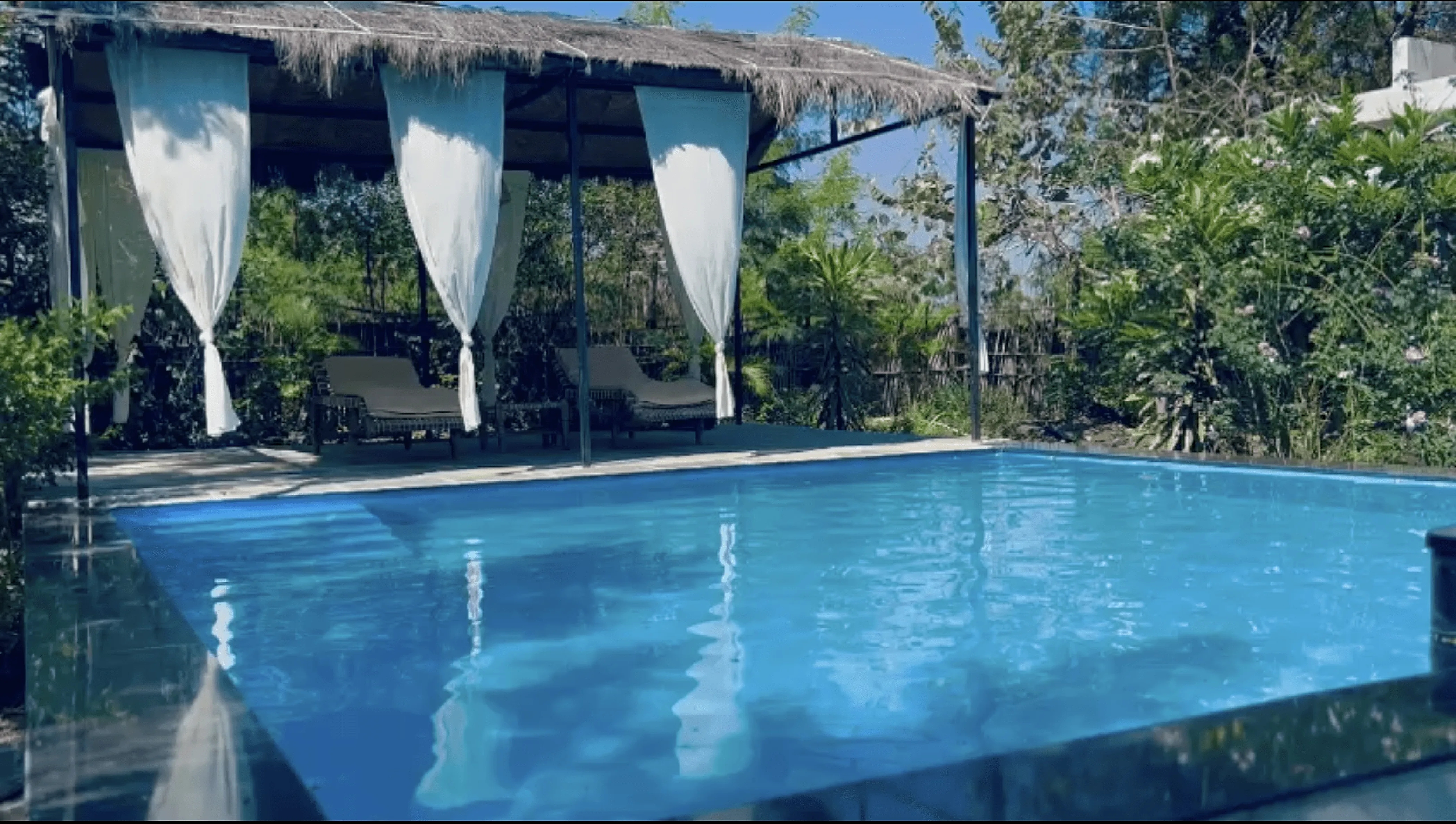 Serene outdoor pool with a thatched-roof cabana, white flowing curtains, and lush tropical greenery in the background at Dhyaana Farms