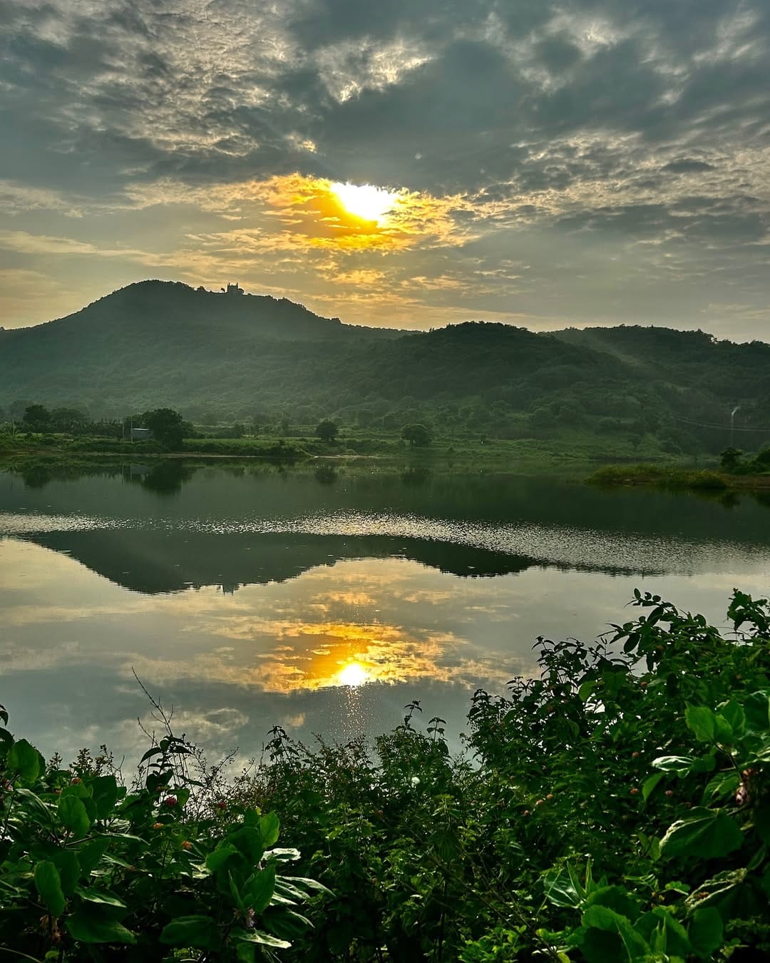Sunset over a serene lake at Dhyaana Farms, reflecting golden light on calm waters with lush green hills in the background.